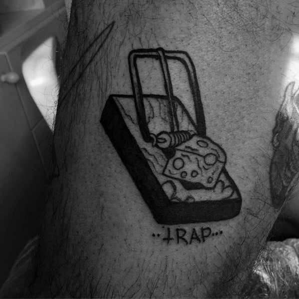 Black and gray tattoo of a mousetrap with a wedge of cheese as bait, featuring bold outlines and the word "TRAP" written underneath.