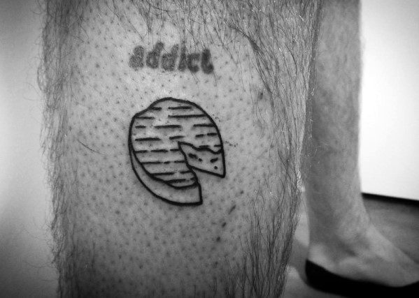 Minimalist black ink tattoo of a round cheese wheel with a wedge cut out, featuring simple line work and subtle shading on the leg.