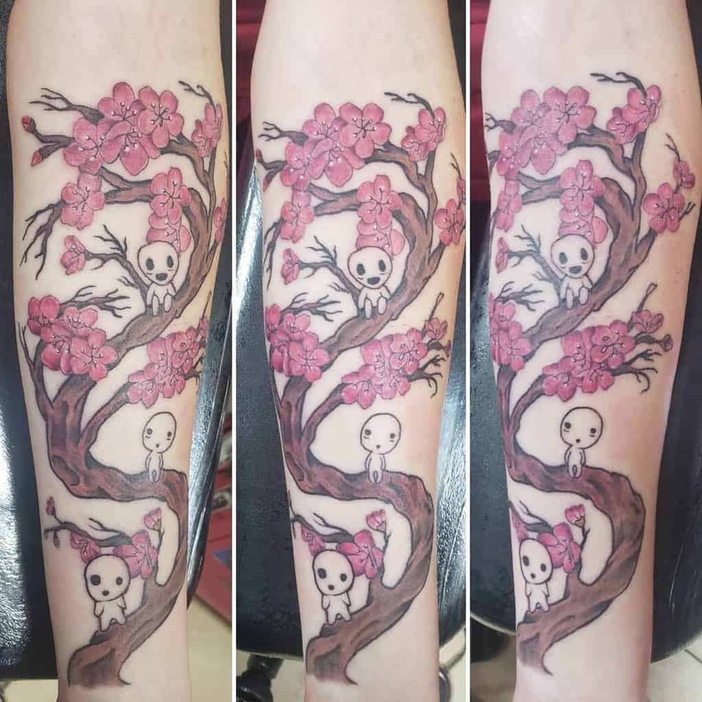 Forearm tattoo featuring a tree with blooming cherry blossoms and kodama spirits sitting on the branches, blending delicate pink shades with fine linework.