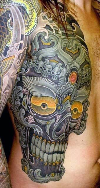 Detailed Tibetan skull tattoo with piercing orange eyes, swirling smoke, and skull motifs on the upper chest and shoulder.