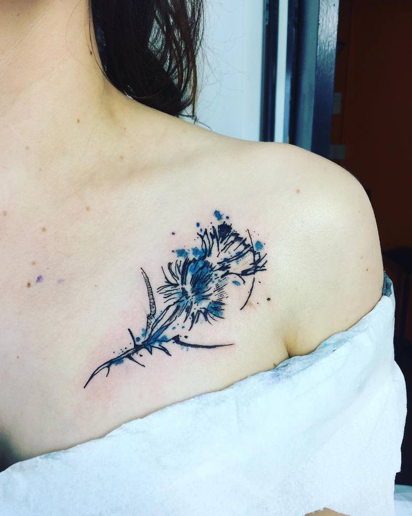 Delicate chest tattoo of a blue thistle with splattered watercolor accents for a subtle artistic touch