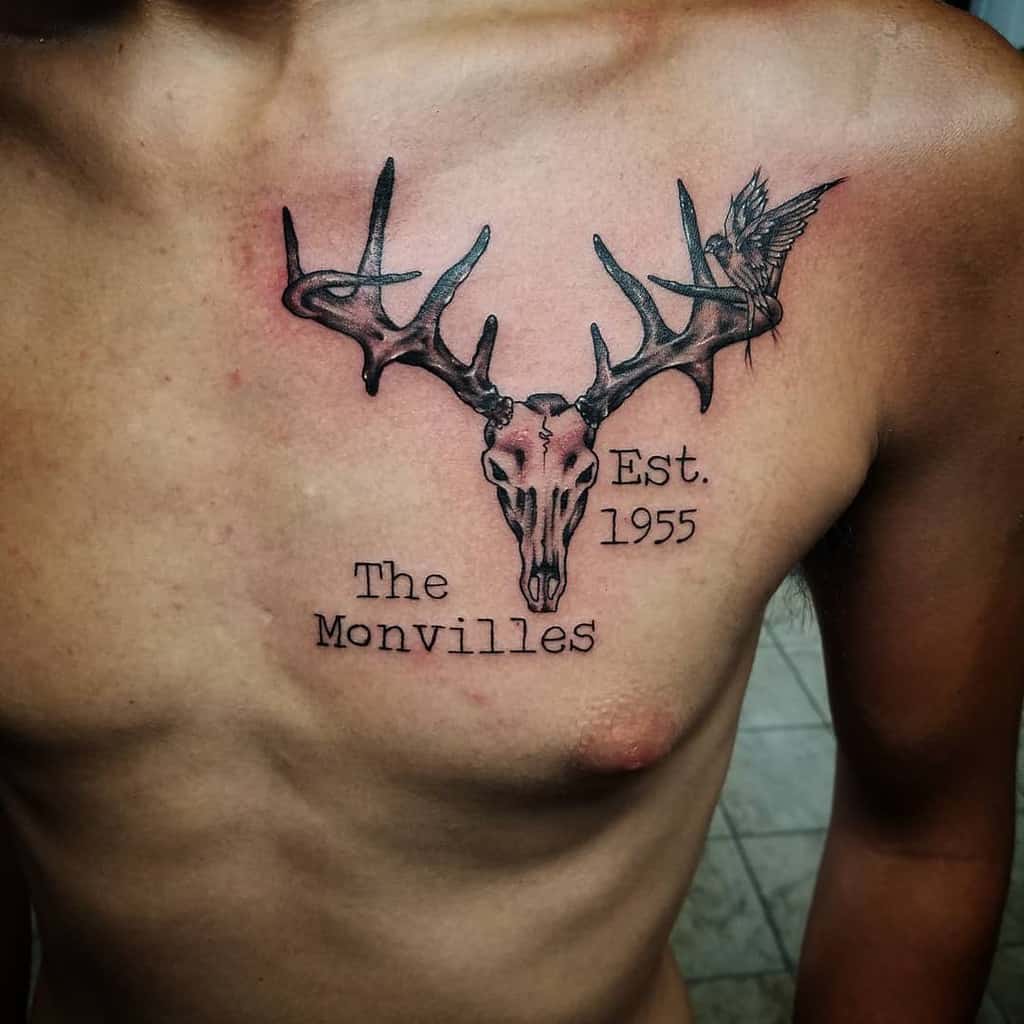 Tattoo of a deer skull with antlers, accompanied by a bird and the text "The Monvilles" and "Est. 1955," etched prominently on the chest
