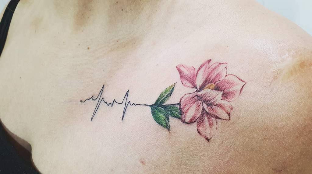 A magnolia flower tattoo in soft pink hues elegantly entwines with a heartbeat line on a shoulder