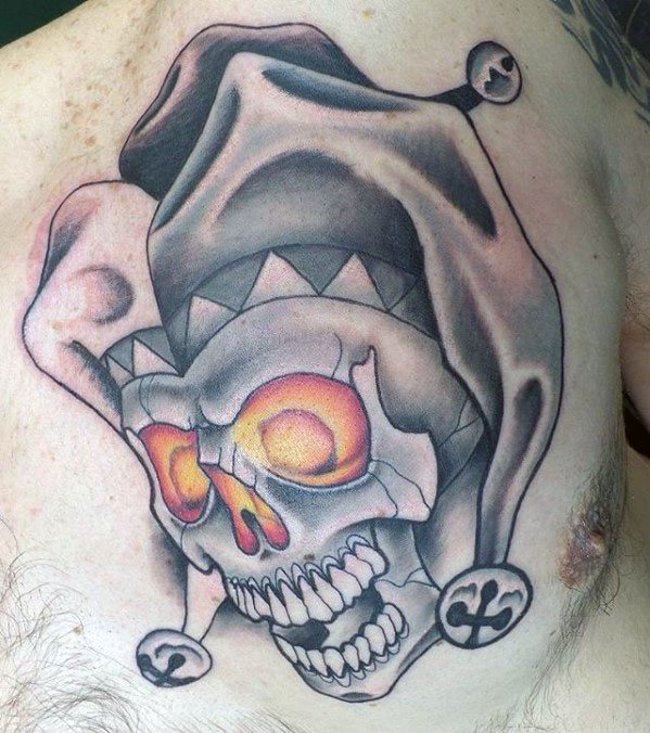 A tattoo of a skull with glowing orange eyes, wearing a jester's hat, designed with bold shading and intricate details on the chest.