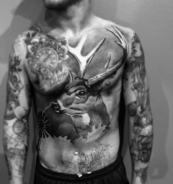 Large black and grey chest and torso tattoo of a realistic buck with antlers, blending into landscape and windmill elements below.