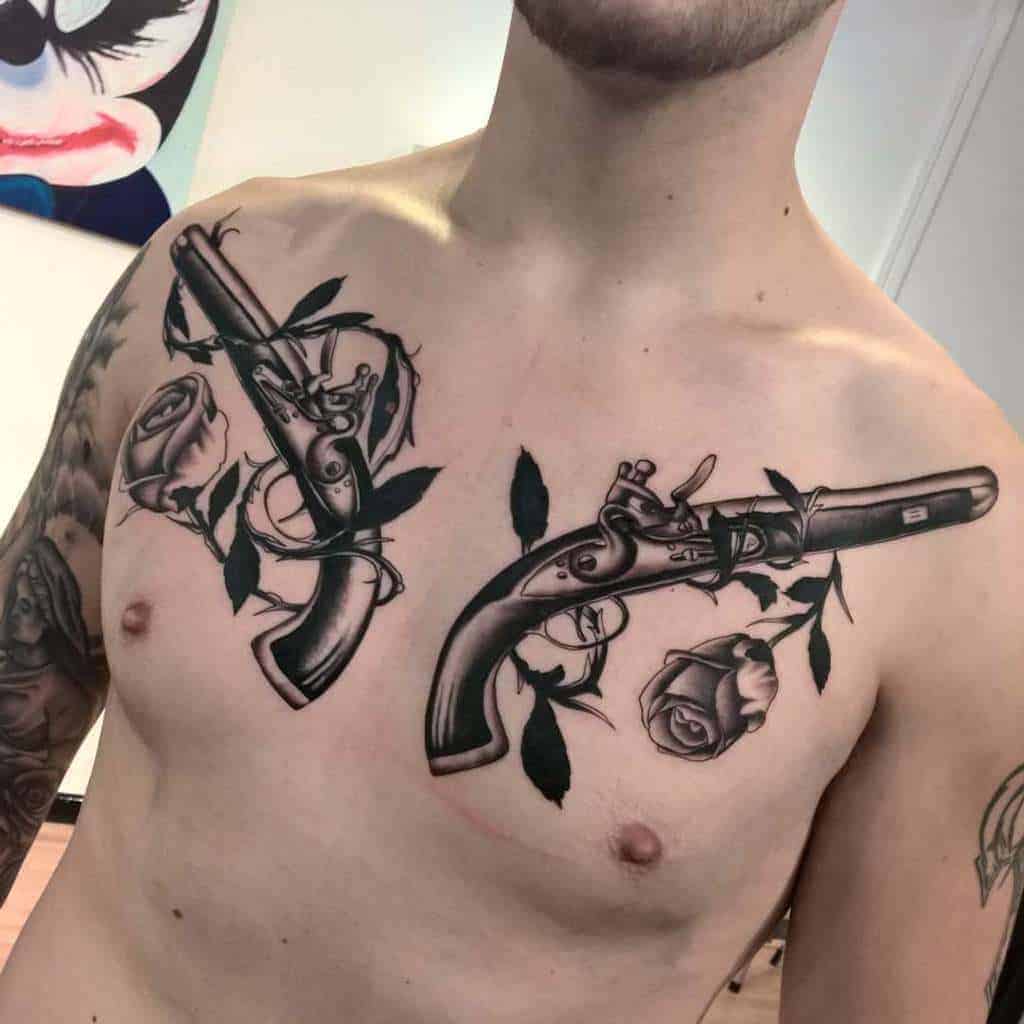 Bold chest tattoo of two antique pistols intertwined with black roses and leaves, creating a symmetrical design