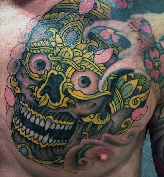 Tibetan skull chest tattoo with gold detailing, pink lotus petals, and smoky background blending into upper arm ink.