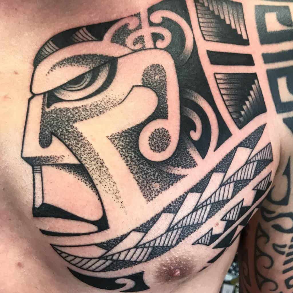 Detailed black and grey Tiki tattoo, featuring a bold mask with geometric patterns, swirling designs, and dot work shading around the face