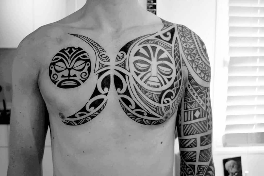 Black and white tribal Tiki tattoo, featuring two detailed Tiki masks, geometric patterns, and swirling designs 