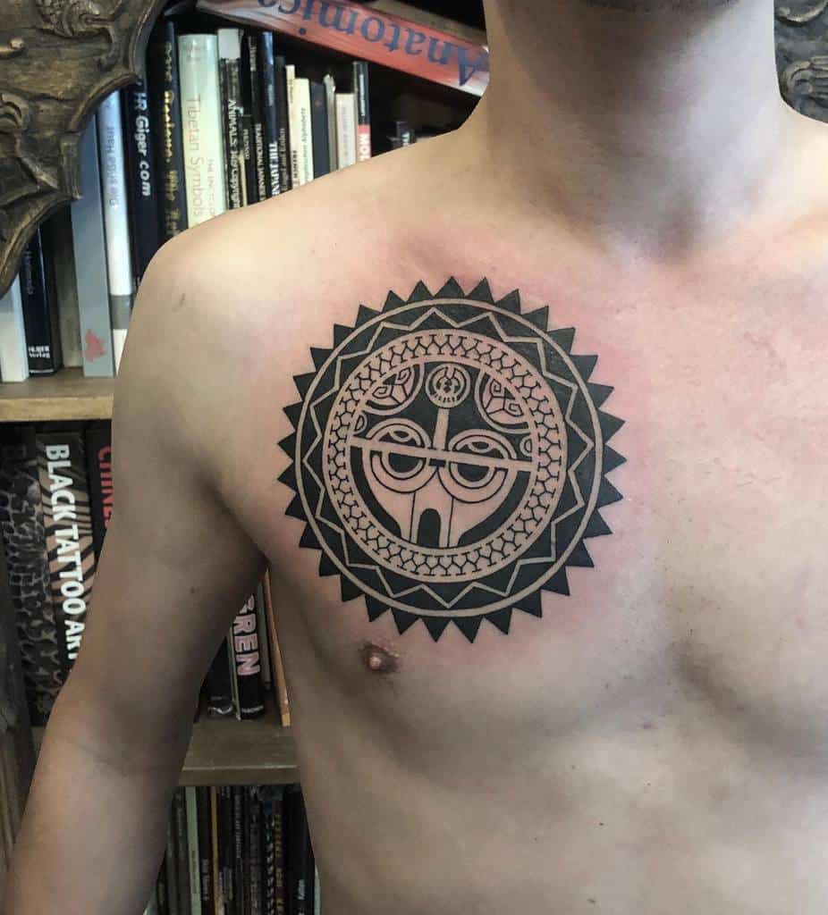 Bold black ink Tiki tattoo on the chest, featuring intricate tribal patterns and a symmetrical circular design inspired by Polynesian art