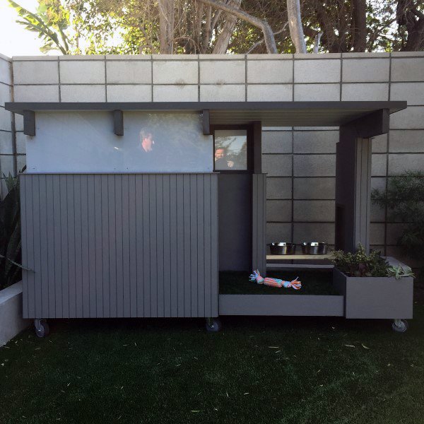 Ultra-modern dog house with a sleek grey design, featuring a shaded area, food bowls, and a toy, set on a grassy surface with a concrete wall in the background