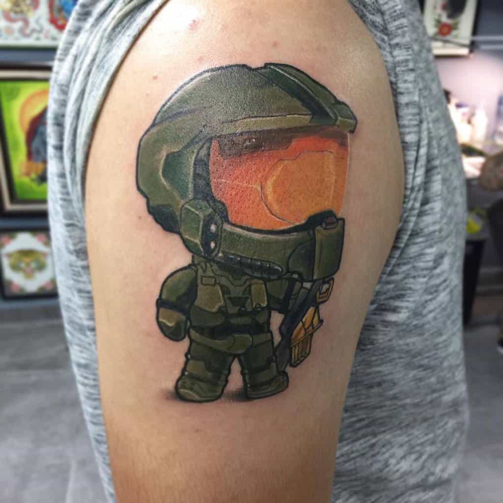 Adorable chibi-style Halo tattoo of Master Chief in green Mjolnir armor, holding a weapon, with bold outlines and vibrant shading on the arm.