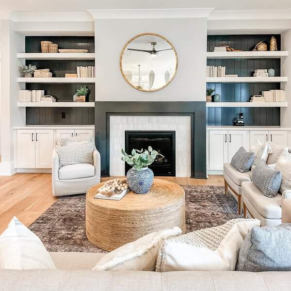 Neutral-toned living room with built-in shelves, a black fireplace, and a round woven coffee table.
