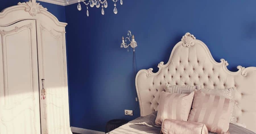 Elegant bedroom with a tufted white headboard, striped pillows, an ornate wardrobe, and a chandelier on a blue wall