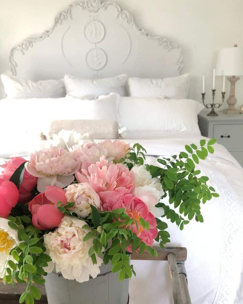 A bright bedroom with white bedding and an ornate headboard, featuring a vase of pink and white flowers in the foreground