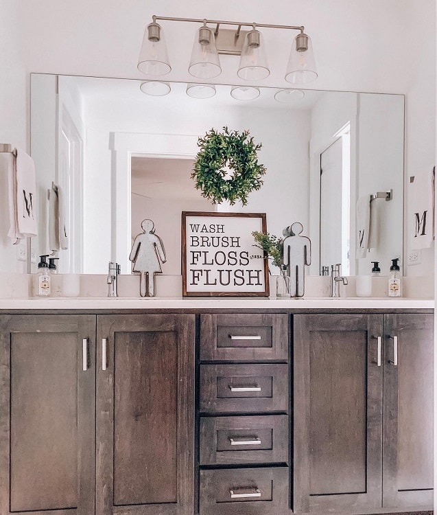 Farmhouse-style modern bathroom with dual sinks, a large mirror, and a leafy wreath above a decorative sign