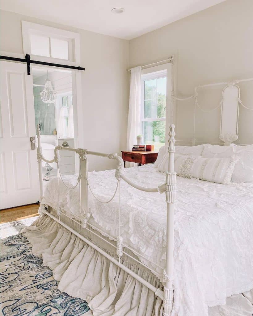 Farmhouse-style bedroom with a white iron bed, ruffled bedding, wooden nightstand, and sliding barn door to the bathroom