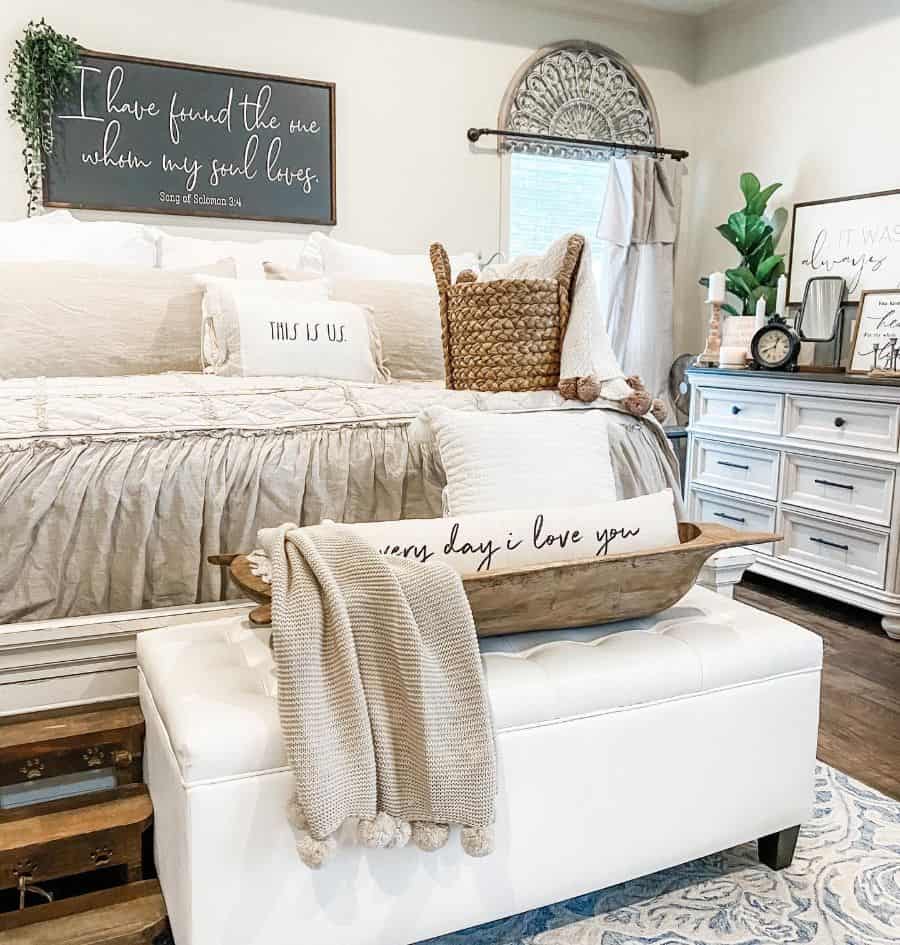 Cozy farmhouse bedroom with a white bed, decorative pillows, a sign above, ottoman, and woven accents for warmth