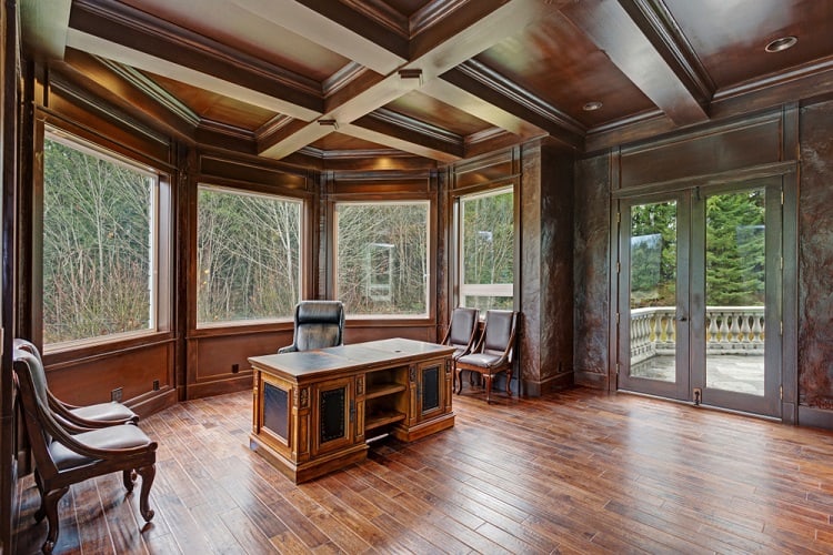 A chic home office featuring a coffered ceiling, rich wood paneling, large windows offering a scenic view, and elegant furniture, creating a refined and professional atmosphere