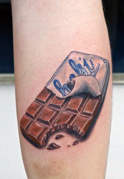 Realistic tattoo of a bitten chocolate bar with a partially unwrapped foil, featuring detailed shading and highlights for a 3D effect on the forearm.