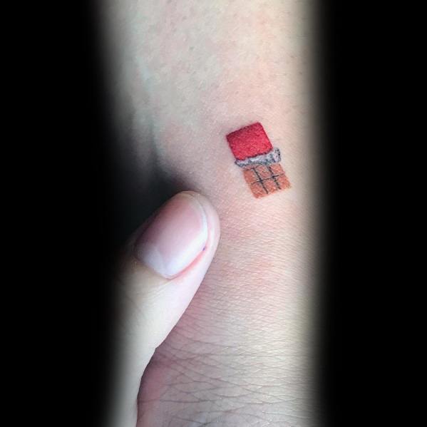 Tiny minimalist tattoo of a partially unwrapped chocolate bar with a red wrapper, placed on the hand with a delicate and simple design.