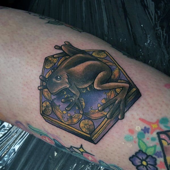 Realistic tattoo of a chocolate frog resting on an ornate hexagonal box, inspired by the magical world, with intricate gold detailing.