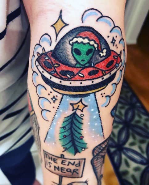 Christmas tattoo of an alien in a Santa hat piloting a UFO, beaming up a decorated Christmas tree, with a sign reading "The End Is Near" for a quirky holiday twist.
