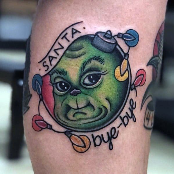 Christmas tattoo of a Grinch-inspired face inside an ornament, wrapped in colorful holiday lights, with a banner reading "Santa bye-bye" for a playful festive twist.