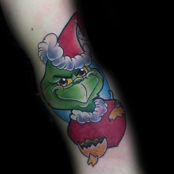 Colorful Christmas tattoo of the Grinch in a Santa hat, smirking while peeking out of a broken red ornament, capturing his mischievous spirit.