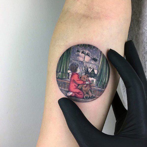 Delicate Christmas tattoo of a child in red pajamas hugging a teddy bear while gazing out a window at Santa’s sleigh flying through the night sky.