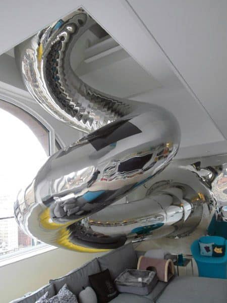 A shiny, metallic slide spirals down from the ceiling in a modern room with large windows and furniture