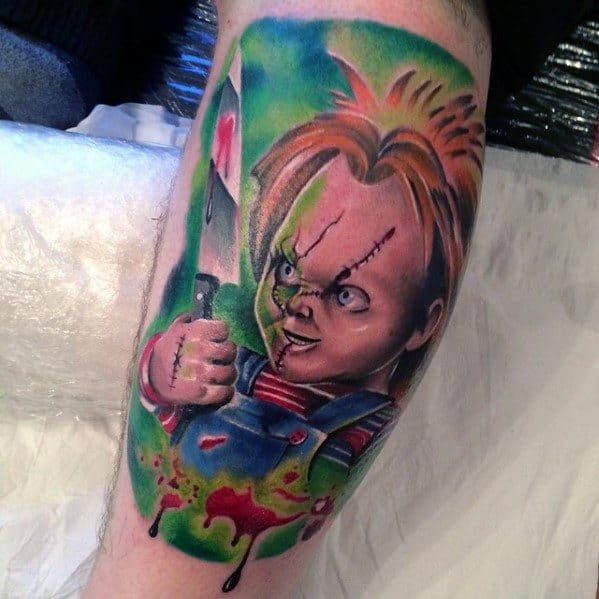 Colorful Chucky tattoo with a sinister expression, holding a bloodied knife, surrounded by a vibrant green background for a striking effect.