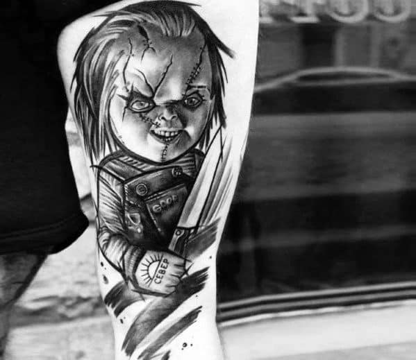 Black and grey Chucky tattoo with bold shading, featuring his sinister grin, stitched face, and a large knife for a dark horror-inspired design.