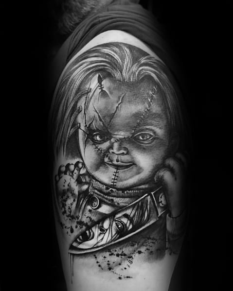 Black and grey Chucky tattoo with eerie shading, featuring his stitched face, sinister grin, and a knife reflecting a terrified face.