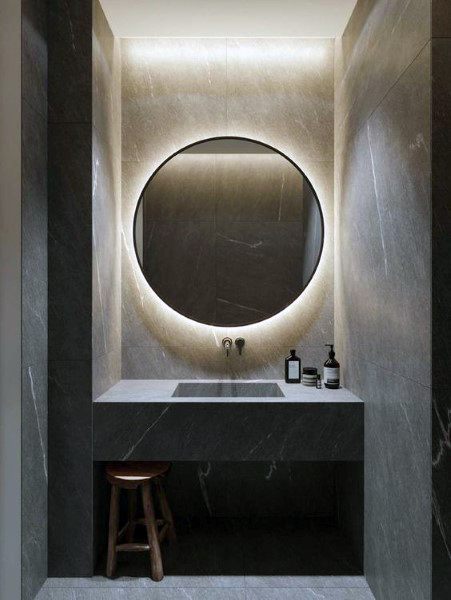 Small bathroom with illuminated round mirror, dark stone walls, and minimalist vanity.