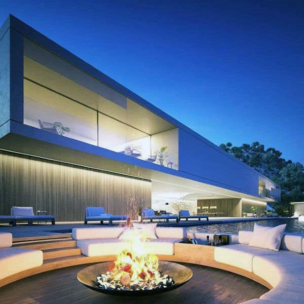 Modern house with large windows overlooking a circular outdoor fire pit surrounded by white seating at dusk