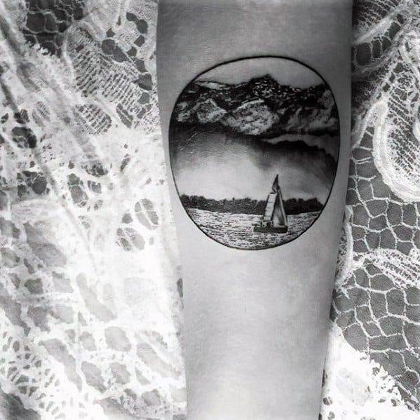 Black and grey lake tattoo with a sailboat on water, framed by distant mountains and trees in a circular window-style design.