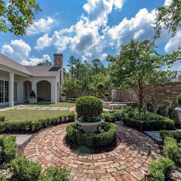Brick patio with circular walkway, lush garden, trees, and a white house under a blue sky