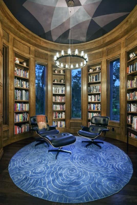 Cozy library featuring wooden shelves, two armchairs, a blue rug, and a chandelier. Large windows frame outdoor trees