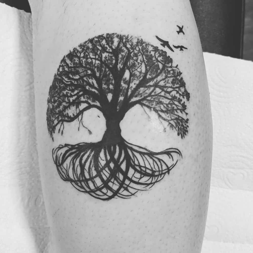The tattoo showcases a detailed tree with expansive roots, complemented by birds flying above, creating a harmonious, nature-inspired design.
