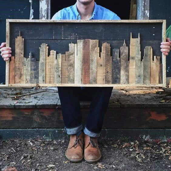 Wooden city skyline art in a rectangular frame held by a person sitting outdoors.