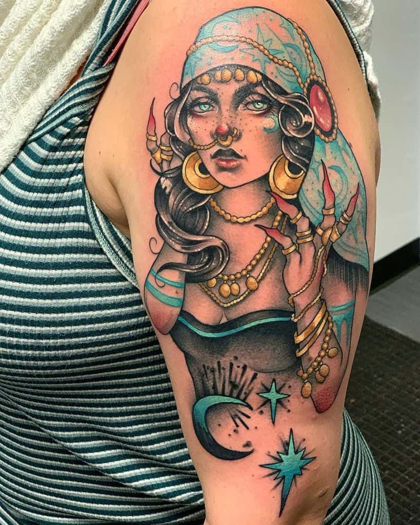 Bold and colorful tattoo of a mystical gypsy woman with glowing eyes, gold jewelry, a flowing headscarf, and celestial details on the upper arm.