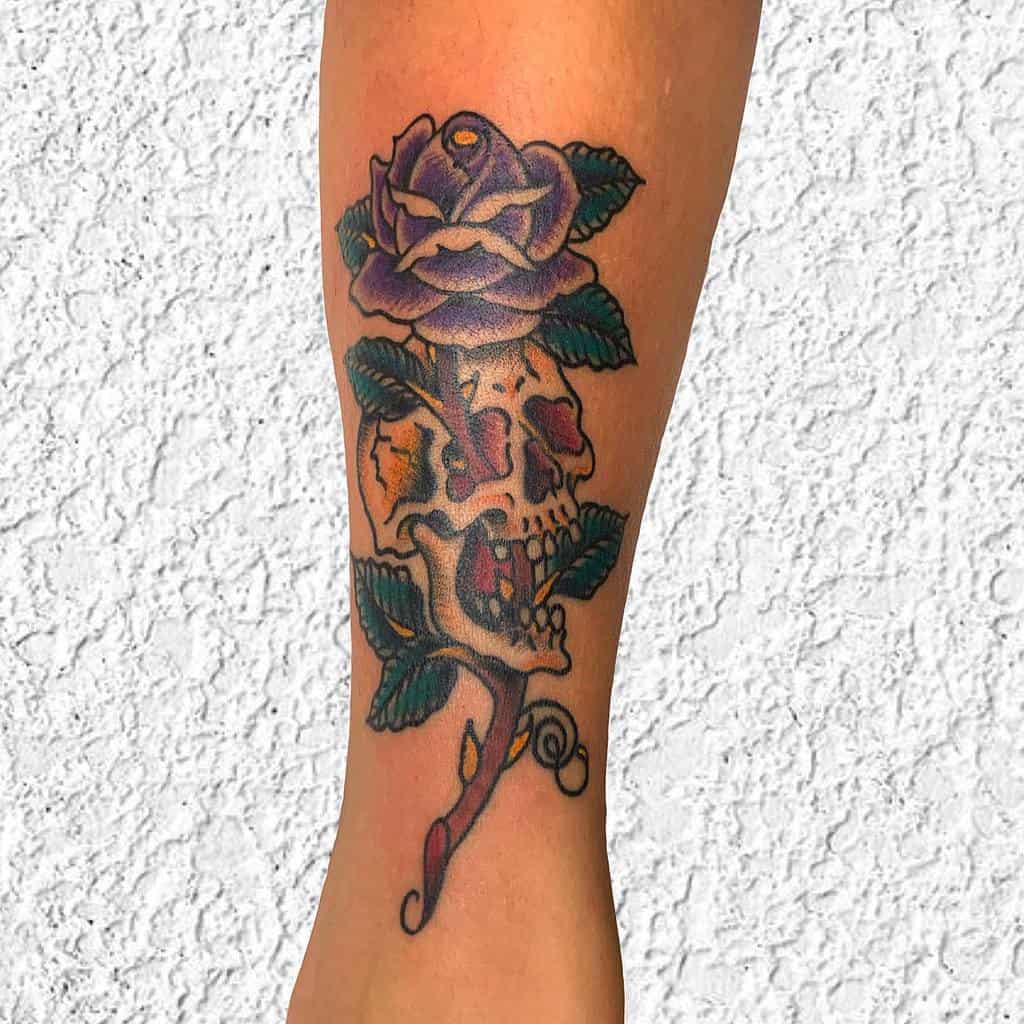 Colorful tattoo of a skull intertwined with a blooming purple rose and green leaves, blending neo-traditional and illustrative styles on the leg.