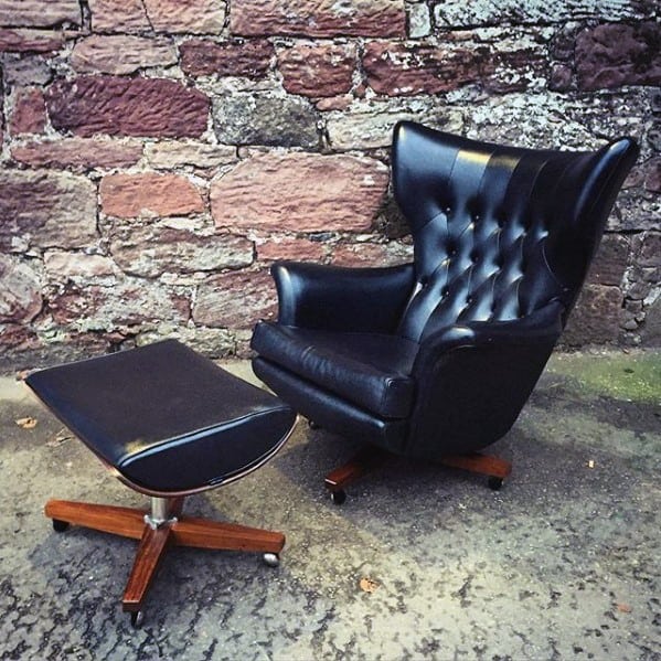 Vintage black leather tufted armchair with matching footrest and wooden base, set against a stone wall backdrop.