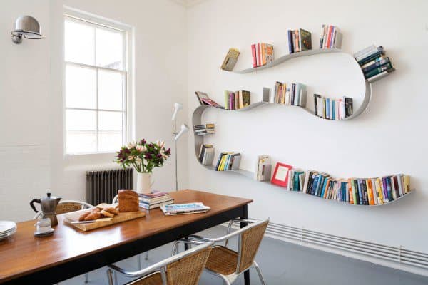 Room with wavy bookshelves, a wooden table with chairs, flowers, books, and bread near a window