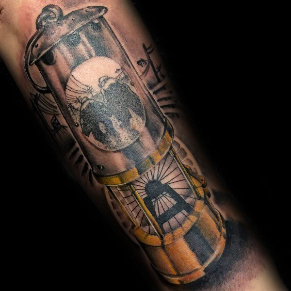 Detailed tattoo of a miner’s lamp with silhouettes of miners and a coal mine structure inside, accented with warm golden tones.