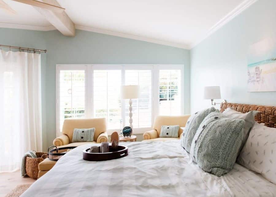 Serene coastal bedroom with light blue walls, wicker headboard, and cozy seating area.