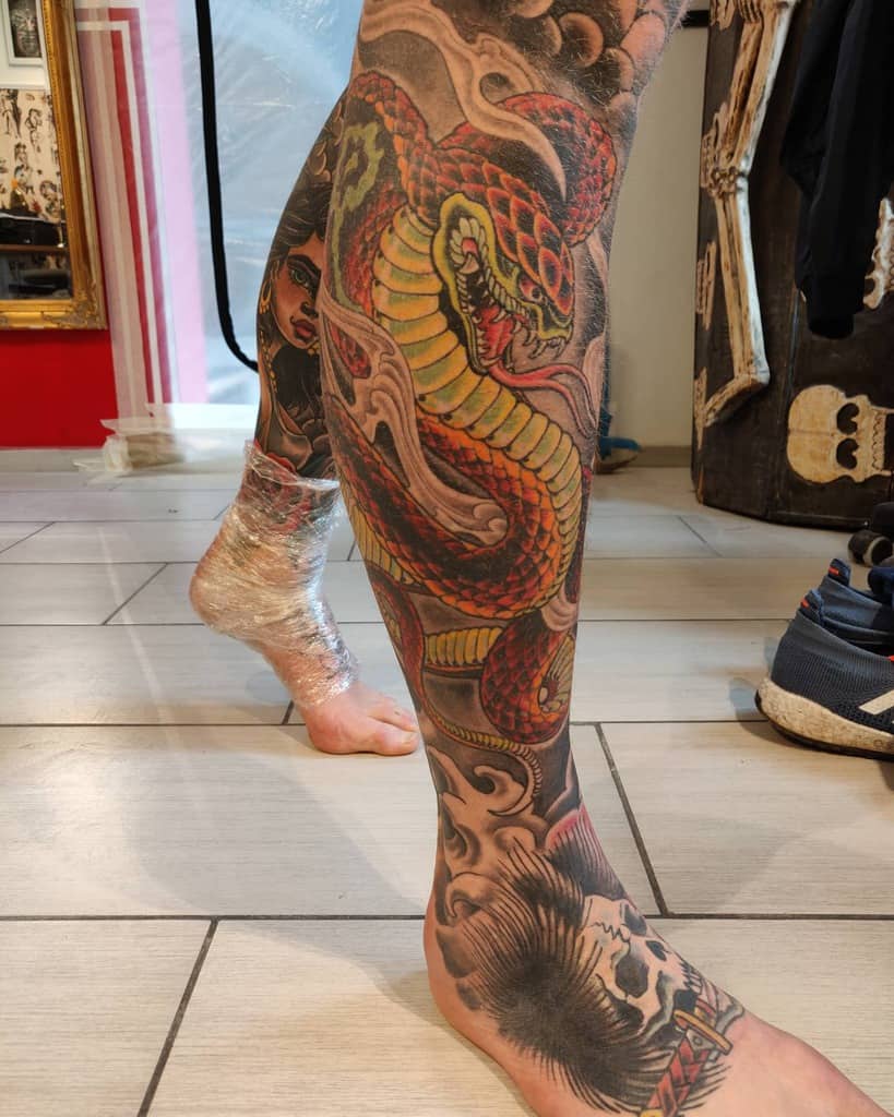 A leg sleeve tattoo featuring a colorful cobra with a background of clouds, along with skull biting a sword