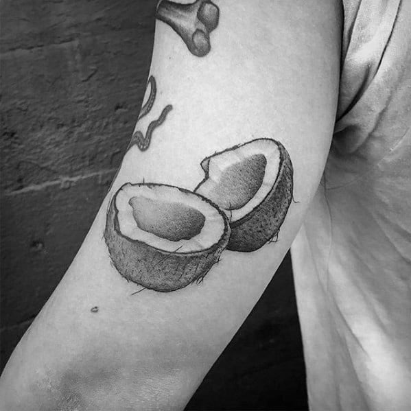 Realistic black and grey tattoo of two coconut halves on the upper arm, showing detailed texture and shading.