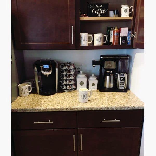 Coffee bar with dark cabinets, granite countertop, coffee machines, mugs, and storage jars.